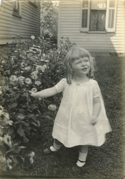 Agnes Moorehead is shown at about 4 years of age. She wears a white dress stands beside a flowering bush.