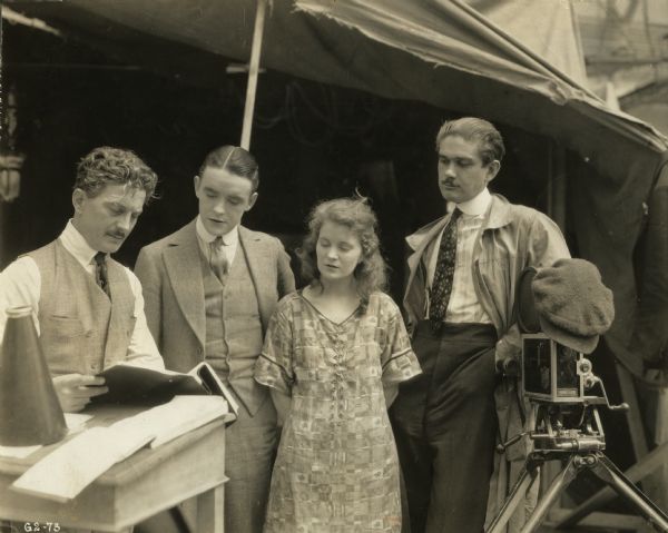 The actors, cameraman, and director discuss the script on location for the silent film "Sunshine Alley" (Goldwyn, 1917). On the right is a Bell & Howell model 2709 camera. On the left is the director's megaphone.<p>Original caption:<p>"Here, for the first time, are Mae Marsh and Robert Harron re-united in a Goldwyn picture. They are the central figures in this photograph and are going over a script with director John Noble. George Hill, Miss Marsh's cameraman, is at the extreme right."