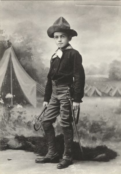 Richard Barthelmess, the future actor, is about 12-years-old in this studio portrait. He is dressed as a soldier, probably one of Teddy Roosevelt's Rough Riders. Behind him is a painted backdrop of a military camp with many tents.