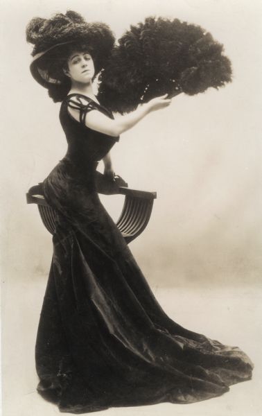 Valeska Suratt's early fame was for her elaborate costumes, wasp waist, ample bosom and hips. She appeared in the 1906 musical <i>The Belle of Mayfair</i> in which she sang "Why do they call me a Gibson Girl?" She wears a long black dress with train and has a large black feather fan and a hat heaped with black feathers.