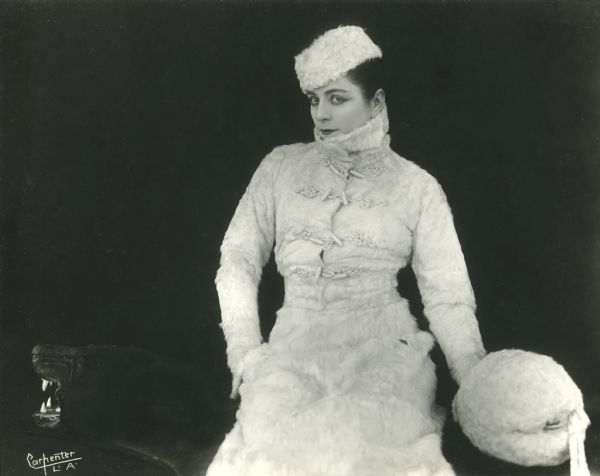 Publicity photograph by Gerald Carpenter of Los Angeles. Three-quarter length portrait of Valeska Suratt wearing a white fur suit, hat, and muffler as she sits atop a black bear rug.