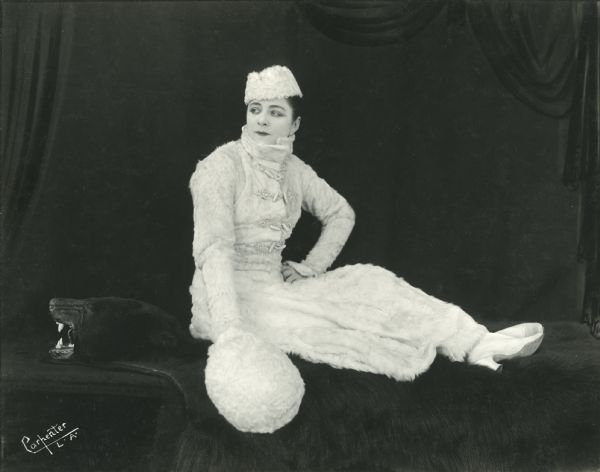 Full-length publicity portrait of Valeska Suratt wearing a white fur suit, hat, and muffler as she sits on a black bear rug.