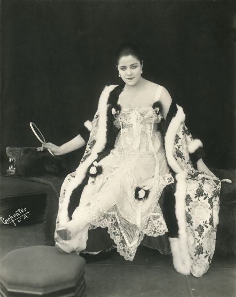 Publicity photograph by Gerald Carpenter of Los Angeles. Valeska Suratt lounges in a dressing gown and net undergarments and holds a hand mirror as she sits atop a black bear rug. The back of the print is covered with biographical notes in pencil, apparently written by a writer for "Photoplay Magazine."