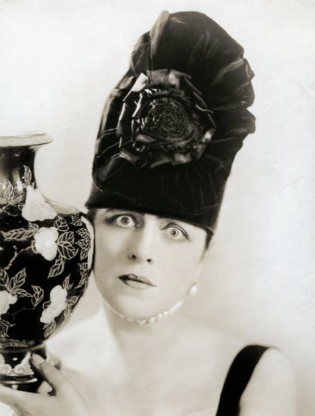 Valeska Suratt, in a tall dark hat, stares intensely at the camera while holding a vase against her cheek.
