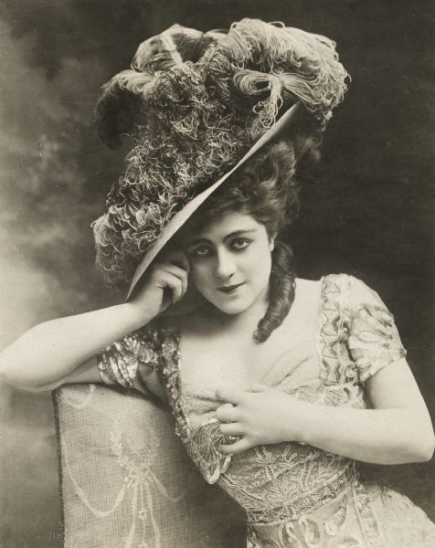 An early waist-up publicity portrait of Valeska Suratt sitting on a chair. She wears an intricate lace dress and a large hat covered with feathers.