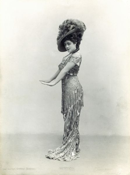 An early full-length publicity portrait of Valeska Suratt in an interesting pose: arms straight out, palms down, as if she were warming by a fire. She wears an intricate lace dress and a large hat covered with feathers.