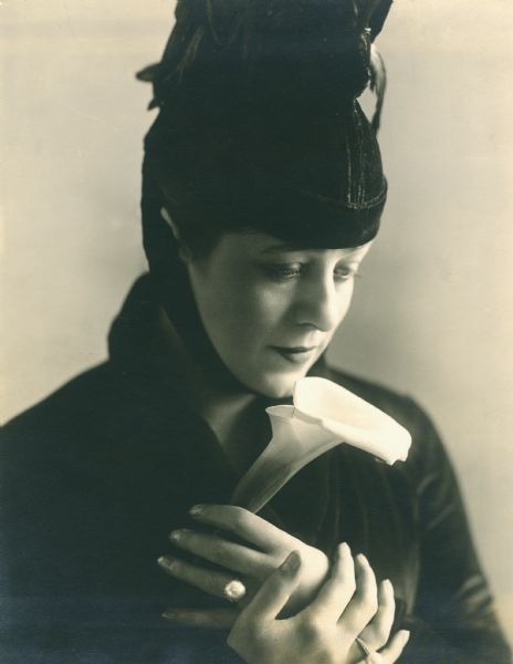 Quarter-length publicity photograph of Valeska Suratt in a dark velvet dress and hat. She holds a calla lilly and wears pearl rings.

The print carries a date stamp, "Sep 30 1918," and stamps for "B.F. Keith's Riverside Theatre" and "to be returned to Photographic & Press Bureau, Palace Theatre Bldg."