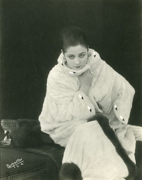Three-quarter length portrait of Suratt wearing a white fur suit as she sits on a black bear rug.