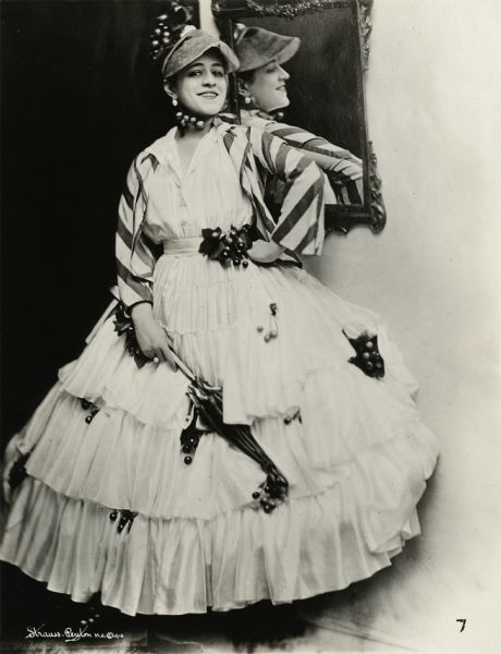 Full-length publicity photograph of Valeska Surrat.

Valeska Surrat, standing beside a mirror, wears a crinoline dress decorated with artificial grapes. She holds a furled parasol and wears a hat and a jacket with wide stripes.