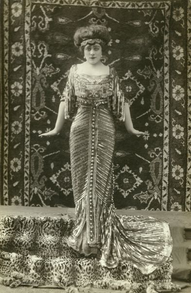 An early full-length publicity portrait of Valeska Suratt posed in the "Egyptian" style. She wears an intricate dress decorated with metallic ribbons and a metal headband. On the wall behind her is an oriental carpet. She stands on a leopard skin.<p>The print is stamped "The Dover Street Studios, 38 Dover Street, Mayfair," a London photographer's studio in business from c. 1906 to c. 1912.