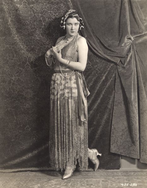 Full-length portrait of Nita Naldi wearing a long pearl necklace and beaded dress. The photograph was probably made by Donald Biddle Keyes as publicity for the 1922 Paramount feature <i>Blood and Sand.</i>