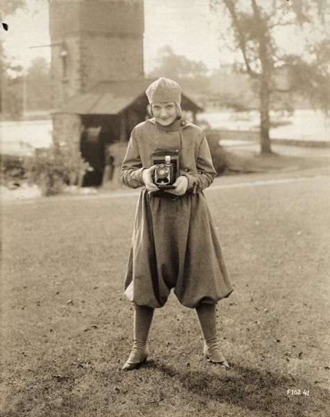 Billie Burke wearing fashionable sportswear (a matching set of bloomers, leggings, and cap) points a Kodak roll film camera at the photographer. She stands in front of a small waterwheel on a pond or river.