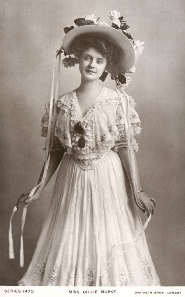 Billie Burke wearing a lovely white dress and large hat decorated with roses in a nearly full length publicity postcard distributed in 1905 for her appearance in <i>The Blue Moon</i> which played at the Lyric Theatre in London, England.

<i>The Play Pictorial</i> No. 82 (1905):
"Miss Billie Burke wears some exceedingly pretty dresses. Her first is of white mousseline de soie, the skirt edged with a number of wee frills above which the mousseline falls in scollops with embroidery at the edge, and a large medallion of roses arranged in each scollop; the bodice displays a charming little pointed cape arrangement which falls over under-sleeves of a succession of tiny frills similar to the bottom of the skirt, and these frills also form the entire bodice. The little cape is decorated with rose embroidery similar to that on the skirt, and she crowns it with a very fascinating hat of white straw with a wreath of pink roses and foliage arranged carelessly upon the brim, pink silk strings completing it."