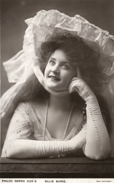 Head and shoulders publicity portrait of Billie Burke resting her cheek on her hand. She wears long white gloves and a large lacy white hat. Published as a Philco Co. postcard.