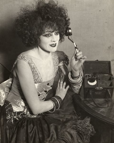 Broadway star Lenore Ulric, dressed and made-up for the stage, listens to an early portable radio receiver.