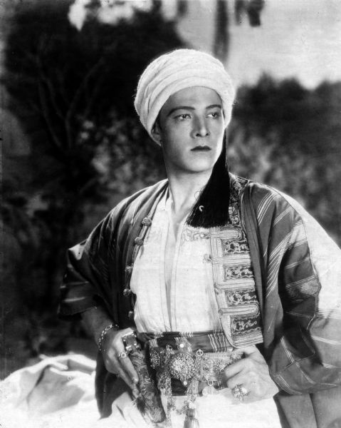 Rudolph Valentino stands with his hands on his hips and wears a white turban in a publicity still from <i>The Son of the Sheik</i> (UA, 1926).
