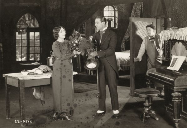 Dorothy Phillips, as Aurora Meredith, receives a bouquet of roses from Rudolph Valentino, playing Juliantino Visconti, from the 1920 Universal production <i>Once to Every Woman,</i> a lost film. They are in a one-room attic apartment with an upright piano. An older man looks on from the doorway.