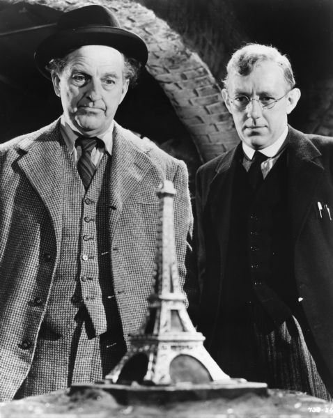 Publicity still of Stanley Holloway (playing Alfred Pendlebury) and Alec Guiness (Henry Holland) contemplating a souvenir Eiffel Tower paperweight in Charles Crichton's <i>The Lavender Hill Mob</i> (1951).