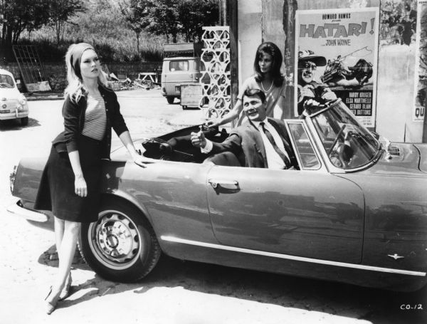 Brigitte Bardot, as Camille Javal, Jack Palance as Jeremy Prokosch, and Giorgia Moll as Francesca Vanini in a scene still from Jean-Luc Godard's <i>Le mépris (Contempt,</i> 1963). Palance sits in an Alfa Romeo convertible. Behind is a poster for the John Wayne film <i>Hatari!.</i>