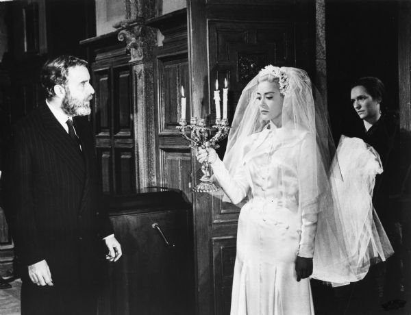 Viridiana (played by Silvia Pinal) wears a bridal gown, her train carried by the housekeeper Ramona (Margarita Lozano) as her uncle Don Jaime (Fernando Rey) looks on in a scene still from Luis Buñuel's 1961 film <i>Viridiana.</i>