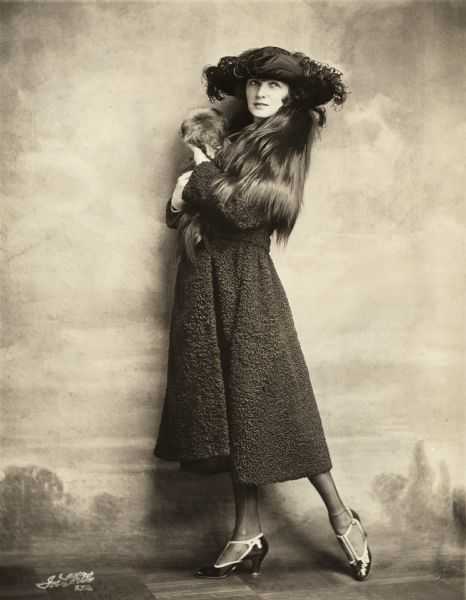 Full-length studio portrait in front of a painted backdrop of Irene Castle in a black Persian lamb coat and hat holding one of her small Brussels Griffon dogs.