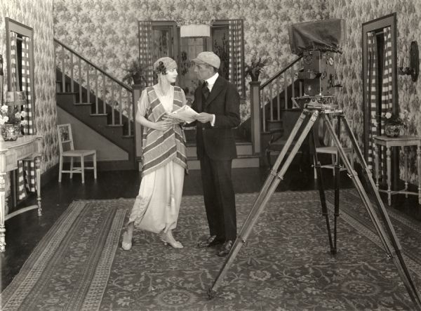 Irene Castle and her director Frank Crane discussing a script on the set of either <i>Vengence is Mine</i> or <i>Stranded in Arcady,</i> two films they made in 1917. In the right foreground is a Pathé 35mm motion picture camera on its tripod.