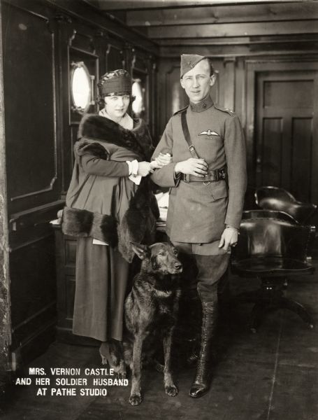 Original caption:<p>"The above picture is the first that has been taken of Mrs. Vernon Castle and her soldier husband since he returned to this country after his strenuous duties as a member of the British Flying Corps at the front in France. Mr. Castle has been awarded the 'Croix de Guerre' in recognition of his distinguished services. He has been visiting his wife at the Pathé Studio where she is starring in motion pictures and the photograph was taken there in a 'set' which is being used in her first feature picture.<p>The dog, it will be noted, is a German police dog. When asked as to how it happens that he is willing to have as pet a dog which is as Teutonic as a dachshund, Mr. Castle informed the interviewer that the canine deserted from the German trenches to the English and was now British in everything except name."<p>This publicity photograph was taken on the set of Irene Castle's silent film "Sylvia of the Secret Service" (Astra Film, 1917).