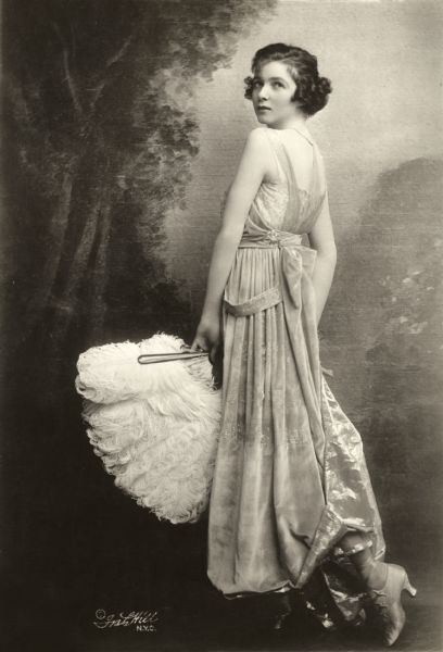 Full-length publicity portrait of Irene Castle posed in a studio in front of a painted backdrop. She looking over her shoulder at the camera and is wearing a light-colored velveteen dress and holding a white ostrich feather fan.