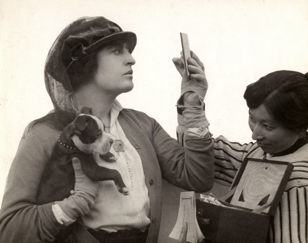 English Shakespearian and Broadway actress Constance Collier checks her makeup in a small mirror held by her maid. Collier holds a puppy.