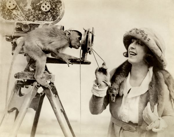 Original title: "This is one of the rare snapshots which a photographer gets only once in a peagreen moon. Dorothy Phillips, star of Allen Holubar's <i>The Soul Seeker</i> is showing her pet simian how a fade out is made with the frontal attachment to the camera. The monk is taking a canny amused interest in the proceeding." Dorothy Phillips, besides being the star of <i>The Soul Seeker,</i> was the wife of the director. The film itself seems to be lost. The camera is a hand-cranked Bell and Howell model 2709, probably belonging to Byron Haskin, the film's cinematographer.