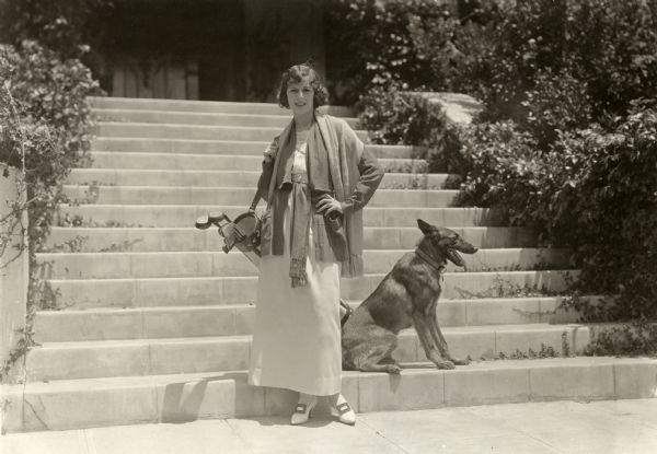 Silent film actress Anita Stewart poses with a golf bag slung over her shoulder and a German shepherd dog.