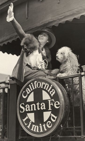 Long time-silent film actress, most notably for Vitagraph and Metro Pictures, Edith Storey, wearing a fox stole, waves good-bye from the platform at the back of the last car of the Santa Fe railroad's Los Angeles to Chicago service, the California Limited. Her dog, probably a Wheaton terrier, is with her. The Santa Fe California Limited logo is prominently shown.