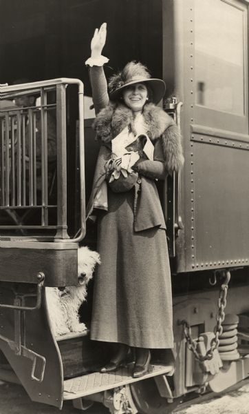 Edith Storey, long-time silent film actress, most notably for Vitagraph and Metro Pictures, wearing a fox stole, waves good-bye from the last car of the Santa Fe railroad's Los Angeles to Chicago service, the California Limited. Her dog, probably a Wheaton terrier, is with her.