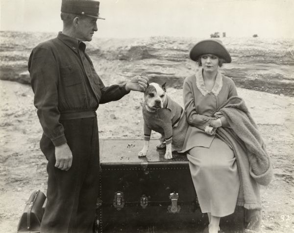 An unidentified actor dressed as a train porter holds a coin out to Blanche Sweet who is sitting on a trunk with her bulldog in a motion picture scene still. The dog wears a sweater.