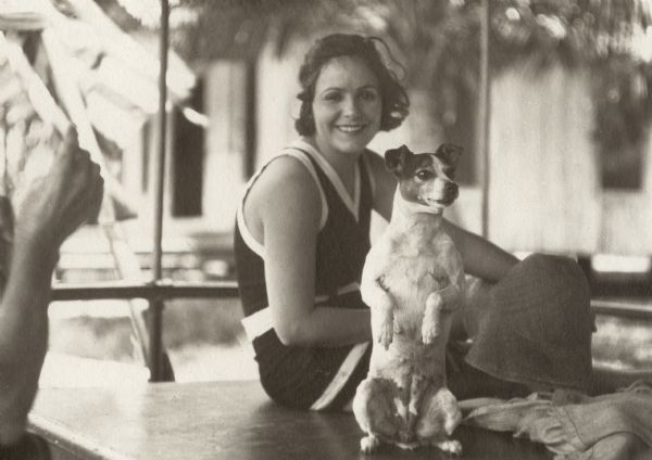 A snapshot of the actress Norma Talmadge wearing a bathing suit. She is sitting on a table behind a rat terrier that is sitting up on its haunches. She was working on a film with the working title of <i>A Broken Barrier</i> which was released in 1919 as <i>The Isle of Conquest</i>.

Original caption:
"Norma Talmadge in Florida where she has been taking scenes for <i>A Broken Barrier</i>.