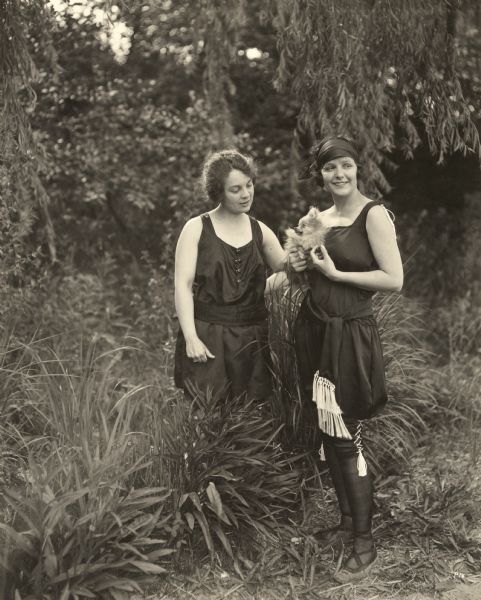 The actress Norma Talmadge is posed outdoors in an athletic outfit, black bloomers with white tassels, holding her Pomeranian dog "Dinky." Next to her is her mother, Margaret Talmadge.
