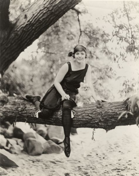 Norma Talmadge sits on a branch of an oak tree with her Pomeranian "Dinky." Talmadge wears black bloomers with white tassels and a black head scarf.