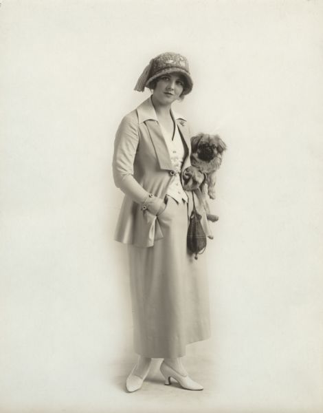 Full-length studio portrait of Triangle films star Olive Thomas wearing a light-colored suit and holding a Pekinese dog under her arm.