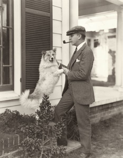 Silent film actor Richard Tucker wears a gray suit and checked cap and has a pipe in his mouth as he pets his dog.  Original caption: "Richard Tucker, Goldwyn player, is very fond of his home and his dog--and it looks as though the dog knew it. Richard hasn't much time to spare these days though as he has just finished in <i>Yellow Men and Gold</i> and is now playing an important role in <i>Remembrance,</i> under the personal direction of Rupert Hughes."