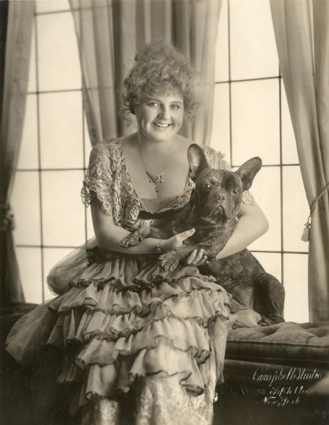 Silent film actress Lillian Walker, in an elaborate ruffled dress, sits at a window holding a black French bulldog.