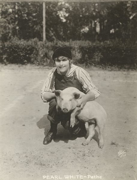Pathé silent film star Pearl White posed with a white pig. White is dressed boyishly, wearing  a black cap, overalls, and striped shirt.