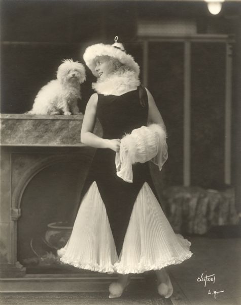Kathlyn Williams wears a white accordion pleated skirt under a black velvet tunic cut into three large triangular sections at the hem. She has a white fur hat and a muff. Her white toy poodle sits on top of a prop fireplace mantel.

Original caption:
"Kathlyn Williams, Morosco star in a powder-puff costume worn to the recent Bachelors' Ball in Pasadena."
