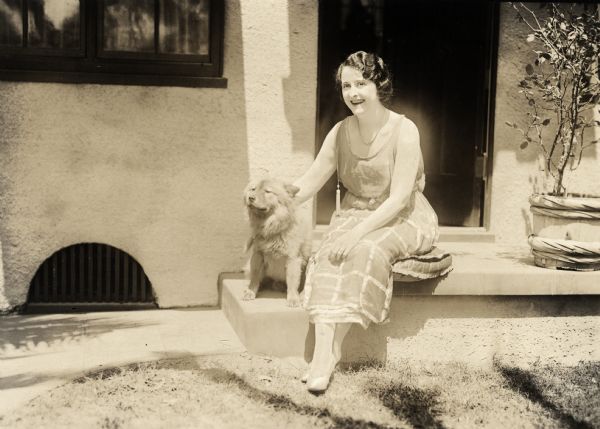 Clara Kimball Young and a chow sitting in the sun on the stoop in front of a stucco house.

Original caption:
"Clara Kimball Young and her prize Chow dog 'Wang.'"