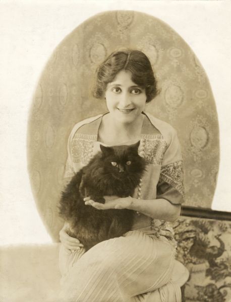 Heavily retouched seated portrait of the silent film star Clara Kimball Young with a black cat on her lap.

Original caption:
"Clara Kimball Young who will soon appear in the World Film production <i>Hearts in Exile</i> with her pet Angora cat."