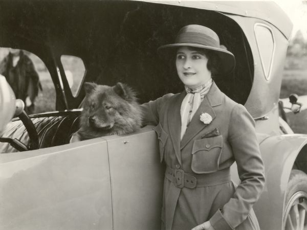 The silent film actress Clara Kimball Young, dressed for a drive in a military-styled coat with patch pockets and a dark straw hat, stands beside a car with a convertible top with her arm around a Chow sitting in the driver's seat.