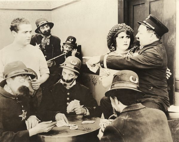 Scene still with Marshall Neilan, in white shirt, and Ruth Roland in a room crowded with police officers from one of the many silent films they made for Kalem in 1912. Most of the officers sport eccentric makeup and costumes.