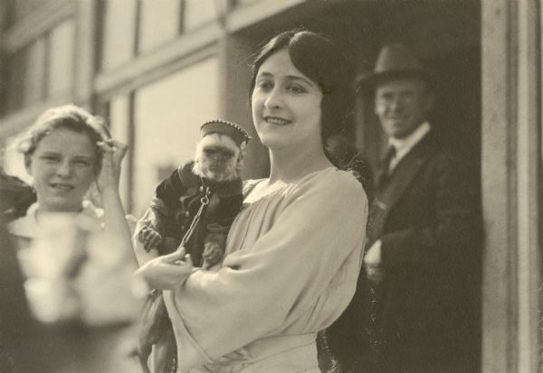 In this snapshot silent film actress Clara Kimball Young holds a white-faced capuchin monkey wearing clothes and a cap. A girl and man are in the background.