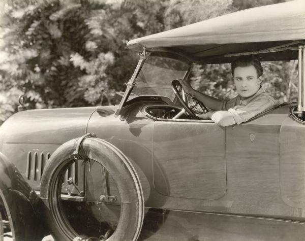 French-born silent film actor Gaston Glass at the wheel of a touring car with a spare tire mounted on the running board. Glass later became an assistant director and was a production manager for the television shows <i>12 O'Clock High, Lost in Space, Batman,</i> and <i>Voyage to the Bottom of the Sea.</i> Original title: "Gaston Glass and his new car, photographed at the Mayer Studio while the hero of <i>Humoresque</i> was working in John M. Stahl's <i>The Song of Life,</i> soon to be released."
