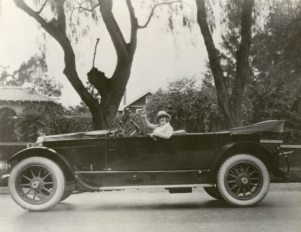 Publicity still of silent film star Mary Miles Minter waves from behind the wheel of her Packard Twin-Six open touring car.

Original caption:
"Mary Miles Minter, the Realart star, in her Packard Twin-Six on her way to work on 'Jerry,' one of the coming Realart productions."
("Jerry" was probably the working title for <i>Don't Call Me Little Girl,</i> a 1921 release in which Minter played a character named Jerry.)