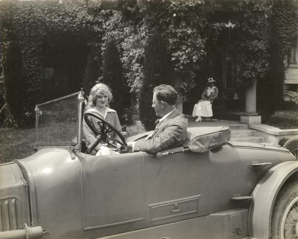 The fifteen-year-old silent film actress Mary Miles Minter stands beside a two-seat roadster automobile looking at the director James Kirkwood sitting at the wheel. A woman sits at the entrance to the house behind them, perhaps Charlotte Shelby, Minter's mother. Minter's career was shadowed by scandal, including an affair with the 42-year-old Kirkwood in 1916 and a connection with the unsolved murder of director William Desmond Taylor in 1922.

Original caption:
"Every morning, James Kirkwood, Miss Minter's director, motors to her home and drives her to the studio."